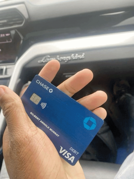 A hand holding a Visa card in the name of Gilbert Arnold Murray, inside a Lamborghini