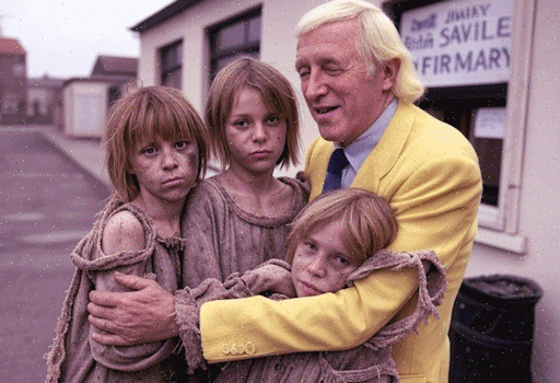 Jimmy Savile hugging three ragged orphan girls, standing outside a building with a Jimmy Savile Infirmary sign in the window
